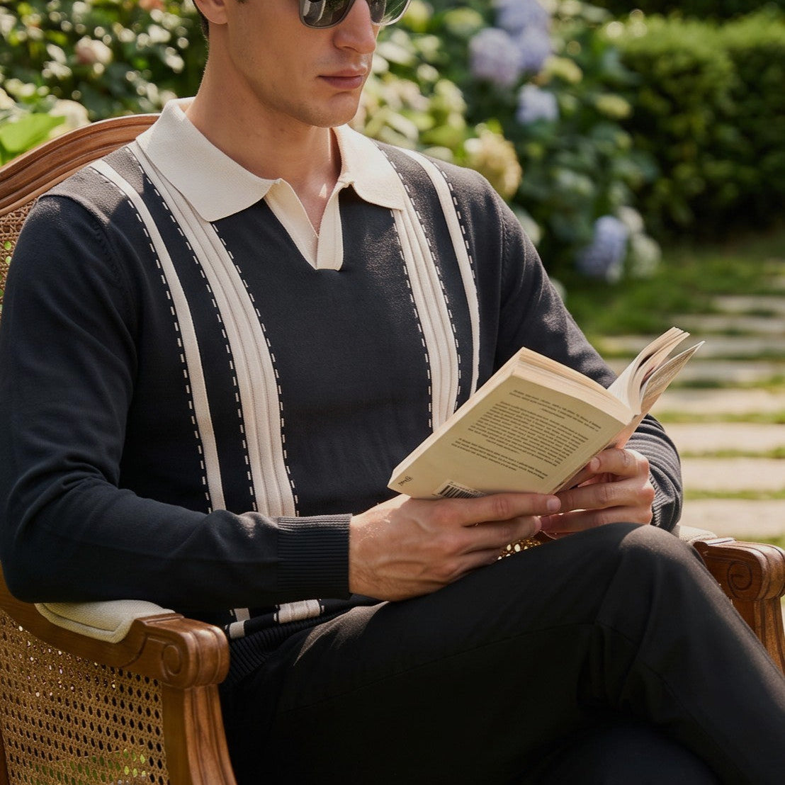 A man wearing aviator sunglasses and a navy knit sweater with cream stripes sits in a wooden garden chair, reading a book.