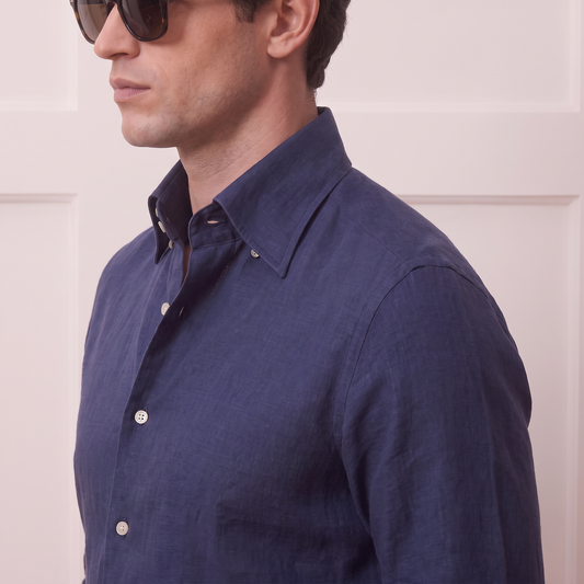 A man in side profile wearing a dark navy blue linen button-down shirt and sunglasses, standing against a light beige paneled wall.