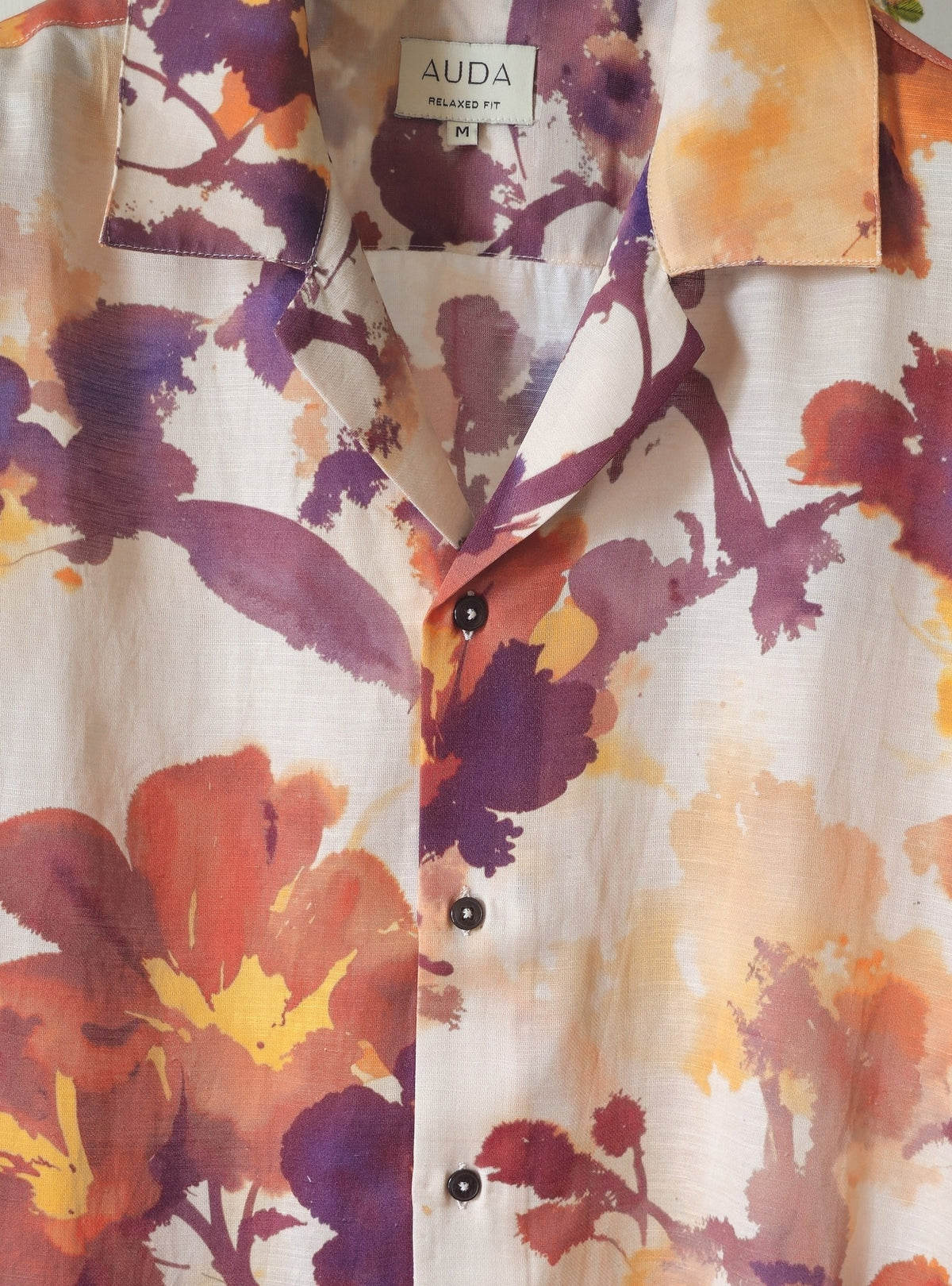A close-up of an AUDA button-up shirt featuring a vibrant watercolor floral print in purple, orange, and yellow on a cream-colored background.