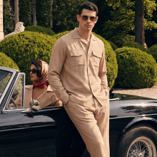 A man in a beige button-up shirt and matching trousers leans against a black vintage convertible car outdoors, with a woman in a matching beige outfit and headscarf seated in the driver's seat. The setting features manicured green hedges and trees.
