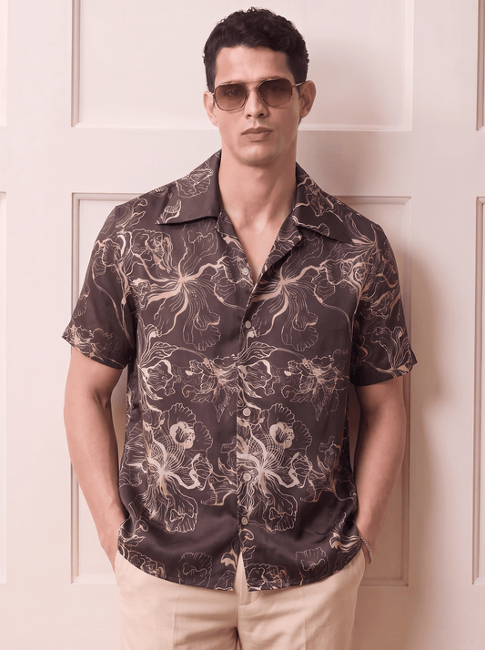 A man wearing a dark brown short-sleeve button-up shirt with a beige and white floral pattern, paired with beige pants and gold-tinted sunglasses, standing in front of a cream-colored paneled door.