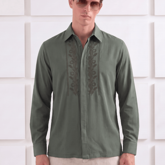 A man wearing a long-sleeve olive green button-up shirt with dark embroidered detailing on the chest, paired with light beige trousers and sunglasses, standing against a white textured wall.