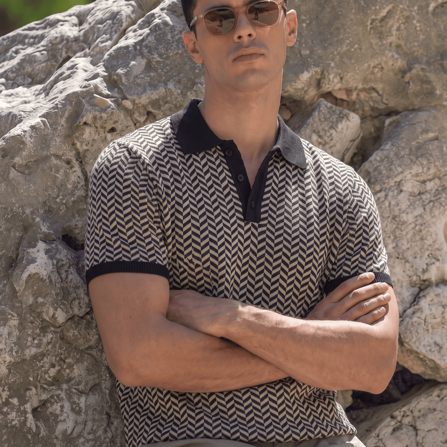 A man wearing a short-sleeve polo shirt with a brown and beige chevron pattern and black collar/cuffs, sitting outdoors against a large rock.
