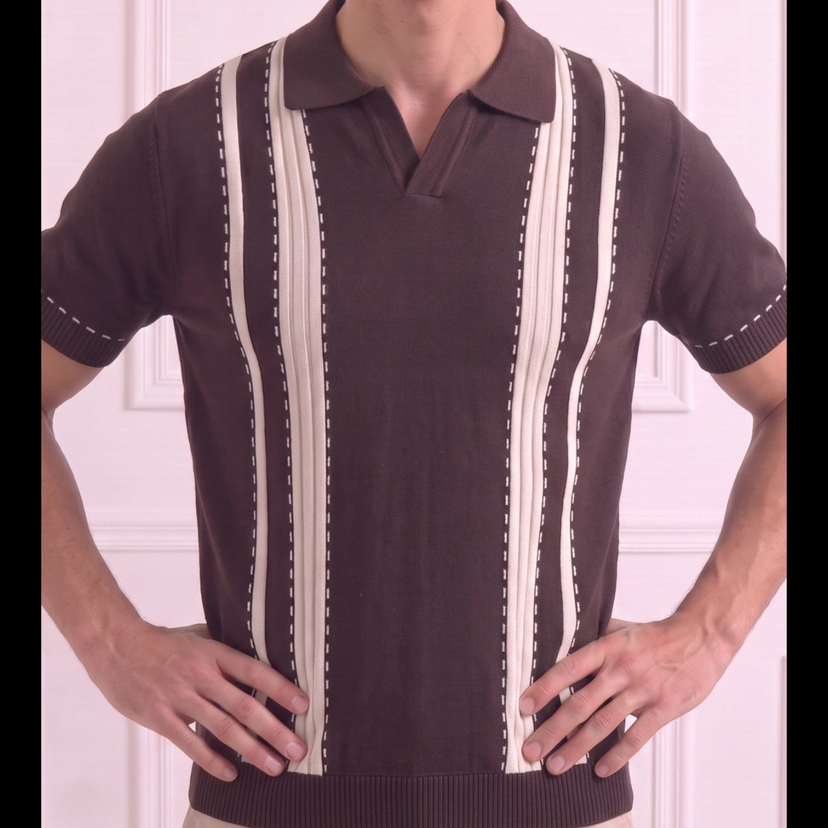 A man wearing a dark brown short-sleeve polo sweater with vertical cream-colored stripes and dotted stitching details, paired with beige pants, standing against a light pink paneled wall.