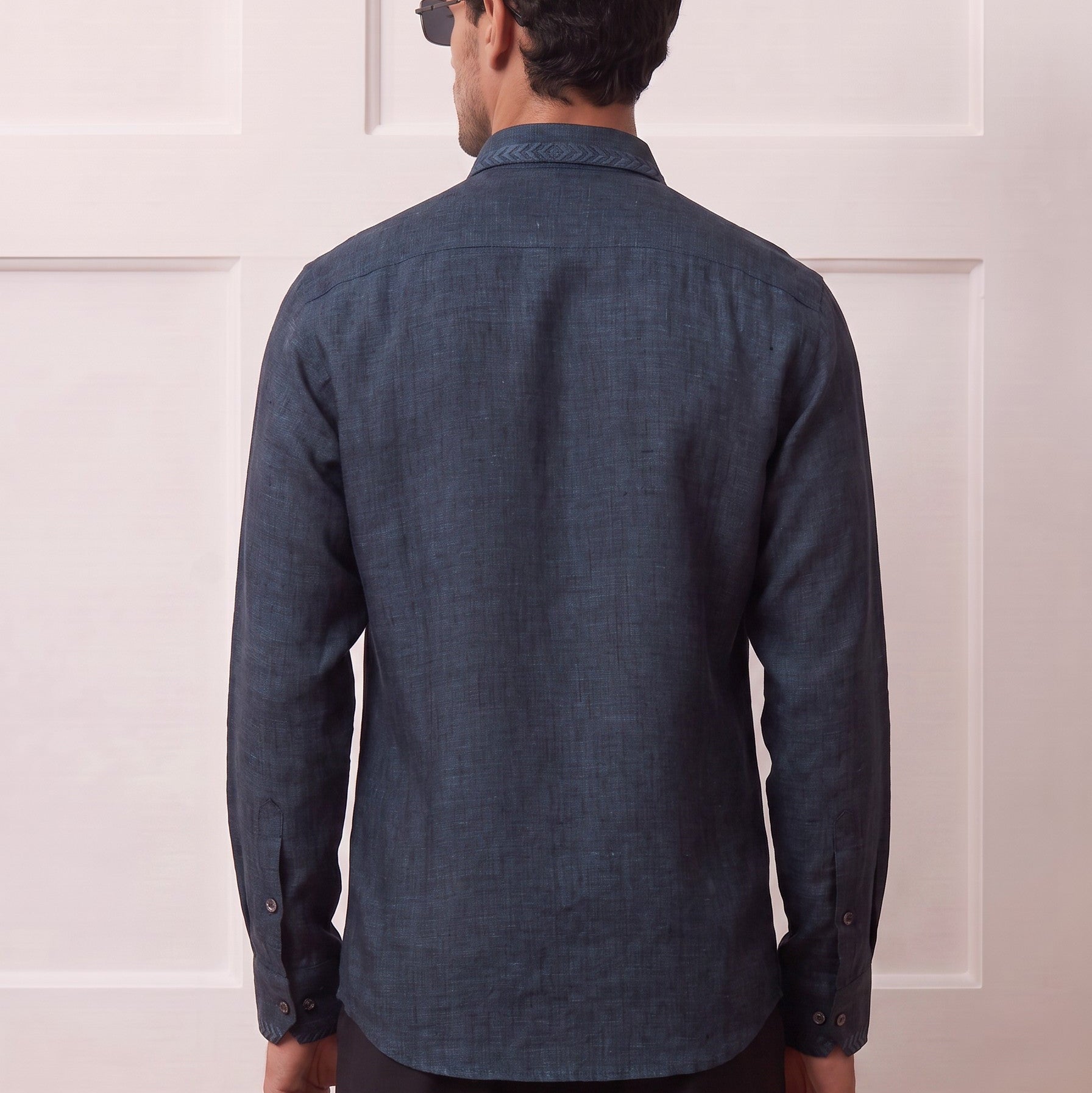 A man viewed from behind wearing a dark blue textured long-sleeve button-up shirt and dark trousers, standing in front of a white paneled wall.