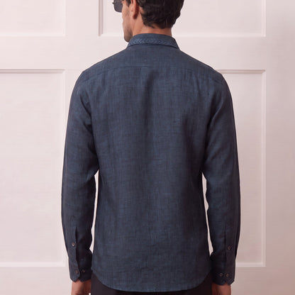 A man viewed from behind wearing a dark blue textured long-sleeve button-up shirt and dark trousers, standing in front of a white paneled wall.