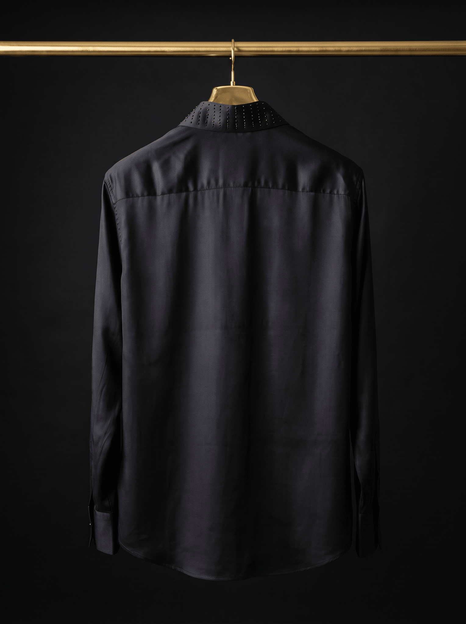 A black long-sleeve button-up shirt hanging on a wooden hanger from a gold clothing rack, shown from the back against a black background.