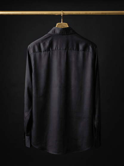 A black long-sleeve button-up shirt hanging on a wooden hanger from a gold clothing rack, shown from the back against a black background.