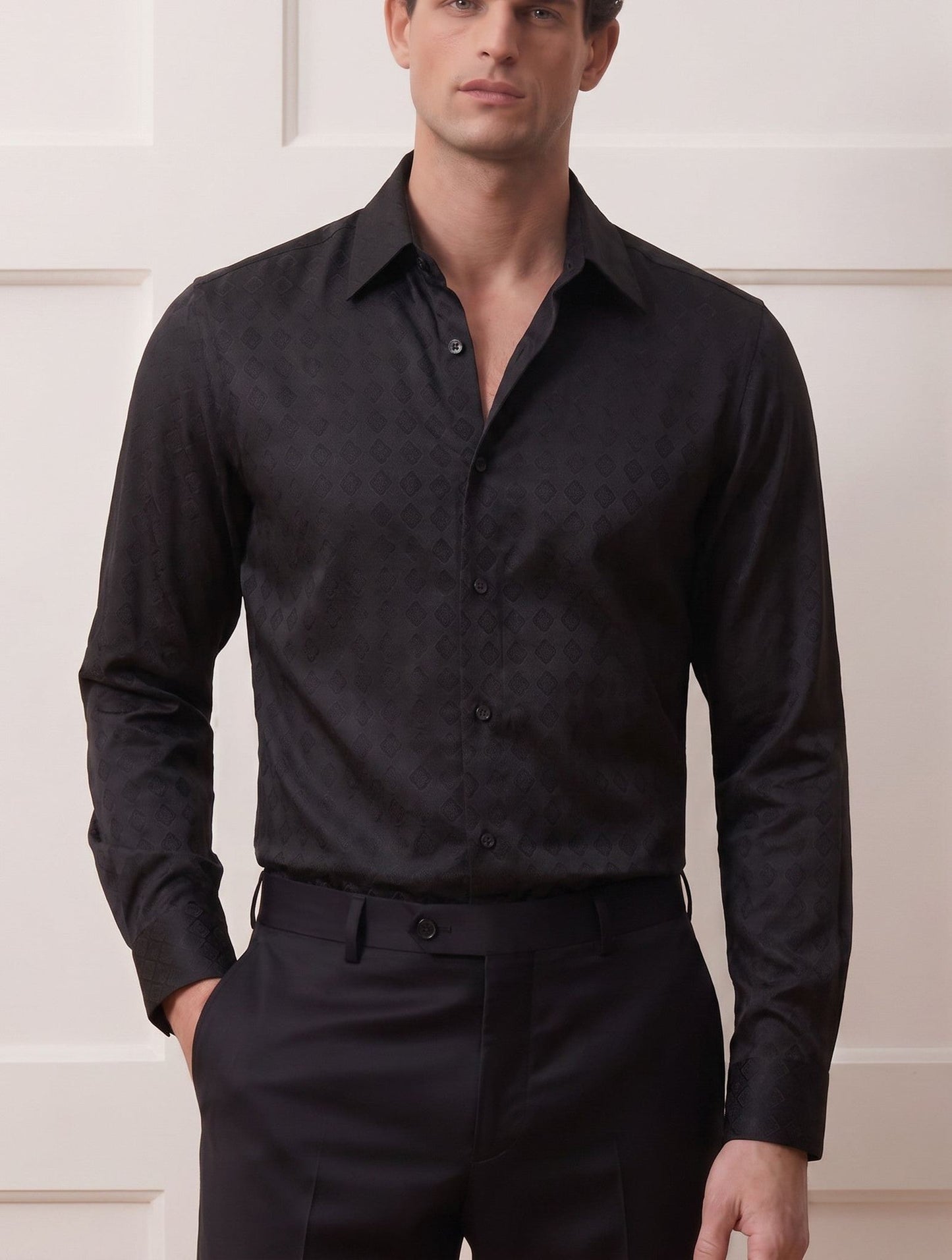 A man wearing a black long-sleeve button-down shirt with a subtle tonal damask pattern, paired with black trousers, standing against a white paneled wall.