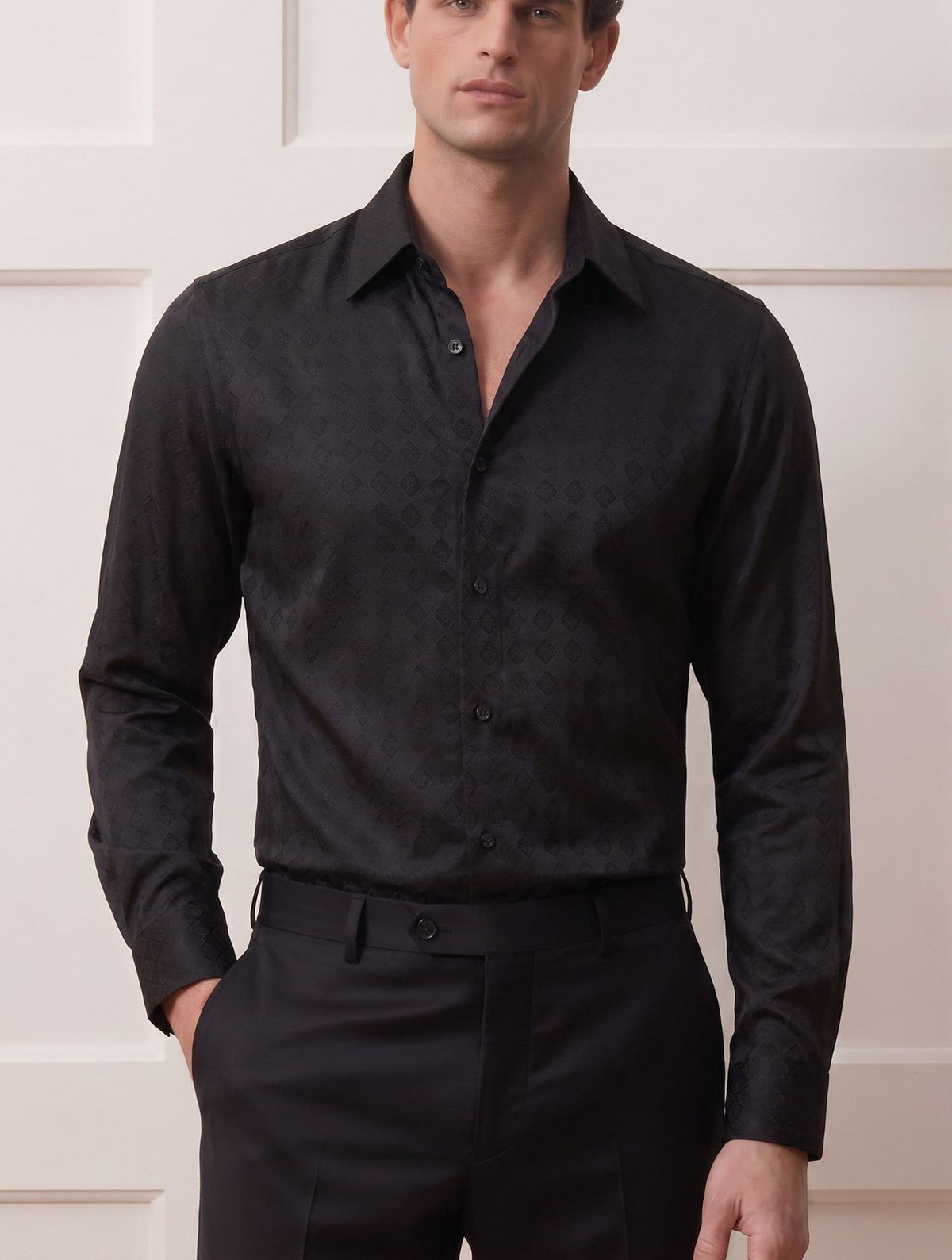 A man wearing a black long-sleeve button-down shirt with a subtle tonal damask pattern, paired with black trousers, standing against a white paneled wall.