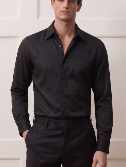 A man wearing a black long-sleeve button-down shirt with a subtle tonal damask pattern, paired with black trousers, standing against a white paneled wall.