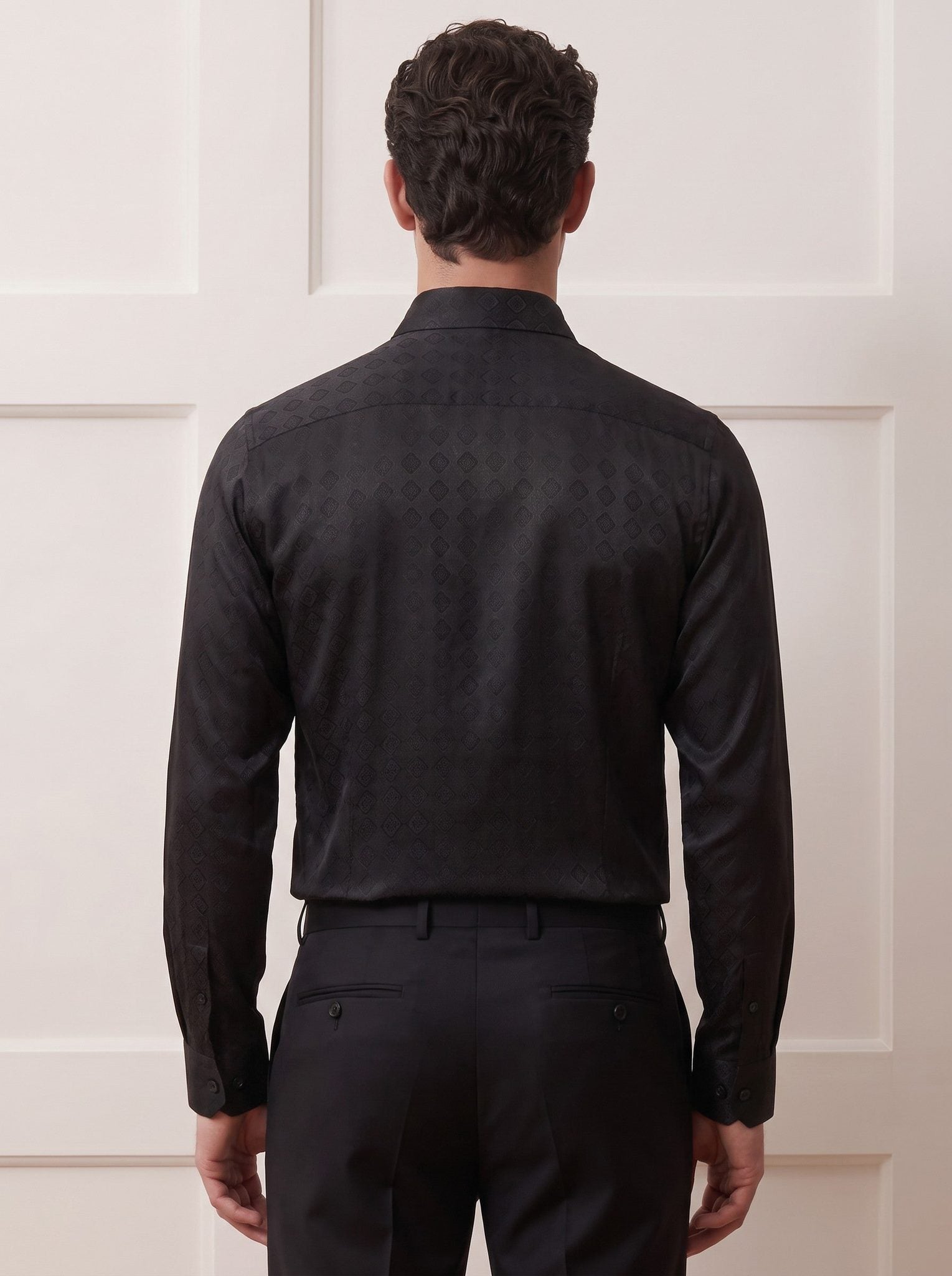 A man viewed from behind wearing a black long-sleeve dress shirt with a subtle tonal jacquard pattern and black trousers, standing against a white paneled wall.