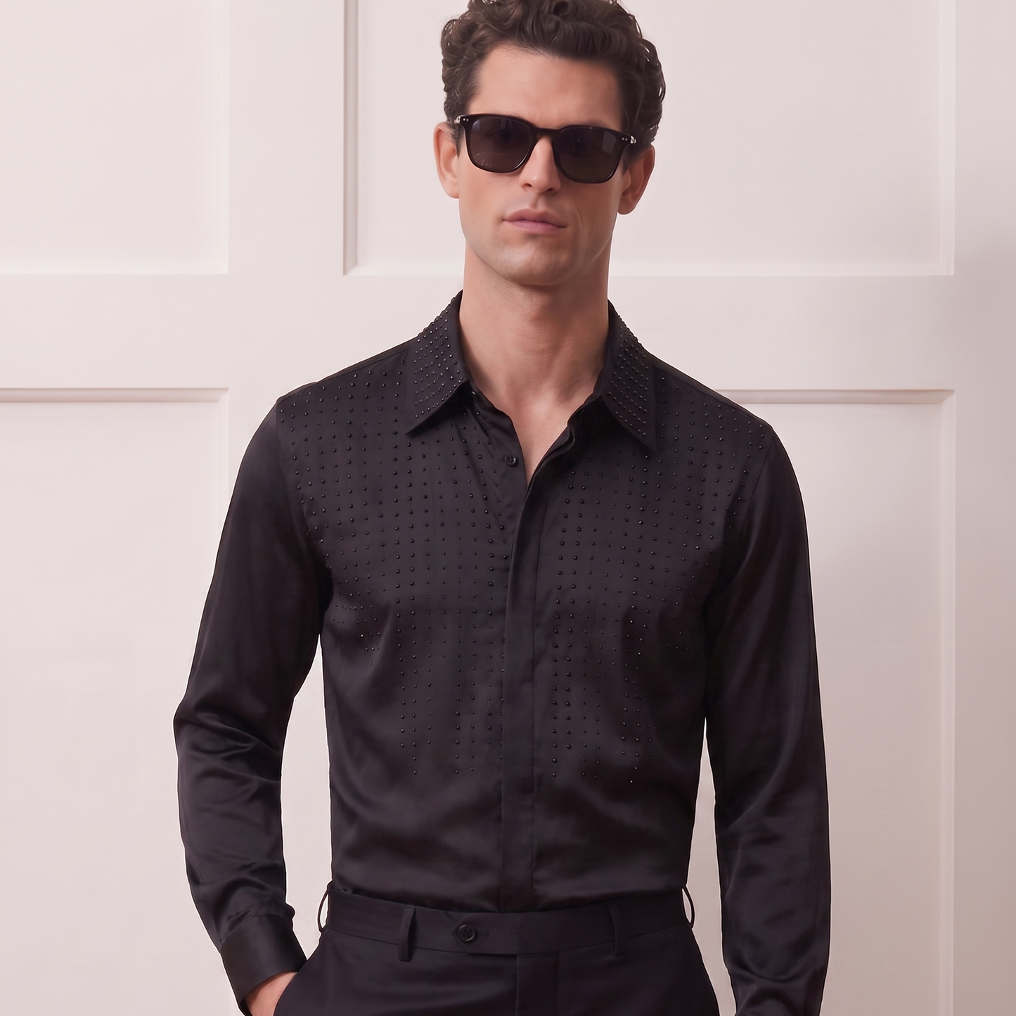 A man wearing a black long-sleeve button-down shirt with a subtle textured pattern, black trousers, and dark sunglasses, standing against a white paneled wall.