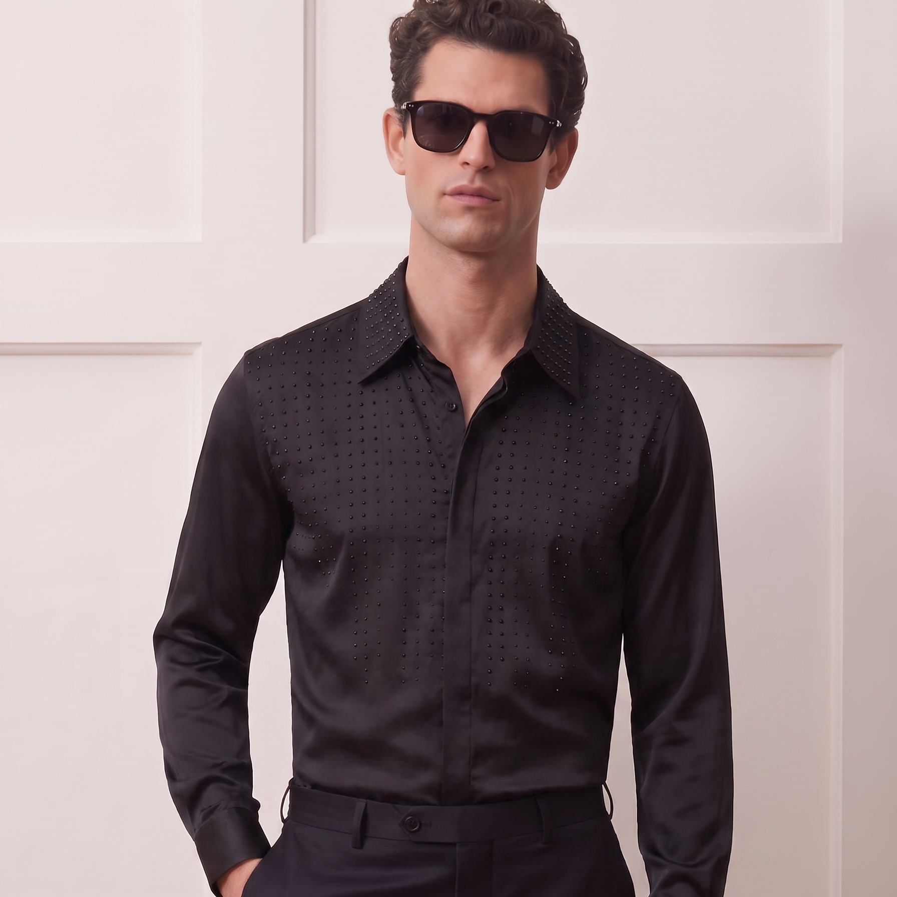 A man wearing a black long-sleeve button-down shirt with a subtle textured pattern, black trousers, and dark sunglasses, standing against a white paneled wall.