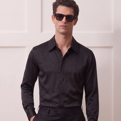 A man wearing a black long-sleeve button-down shirt with a subtle textured pattern, black trousers, and dark sunglasses, standing against a white paneled wall.