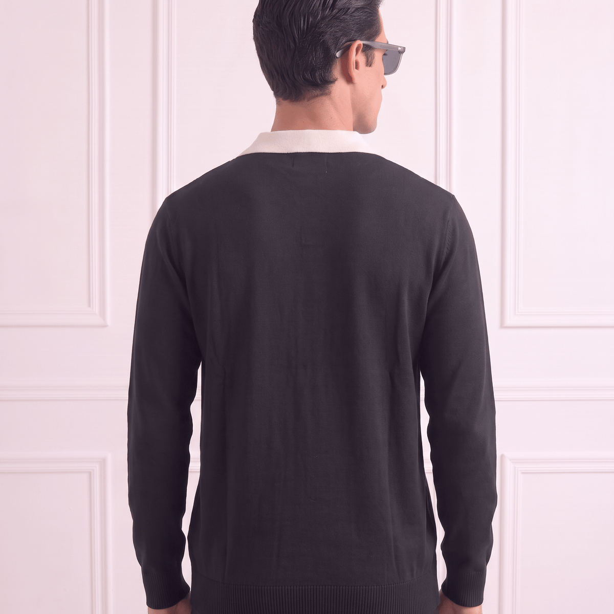 A man viewed from behind wearing a black knit sweater over a white collared shirt, standing against a light pink paneled wall.