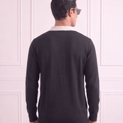 A man viewed from behind wearing a black knit sweater over a white collared shirt, standing against a light pink paneled wall.