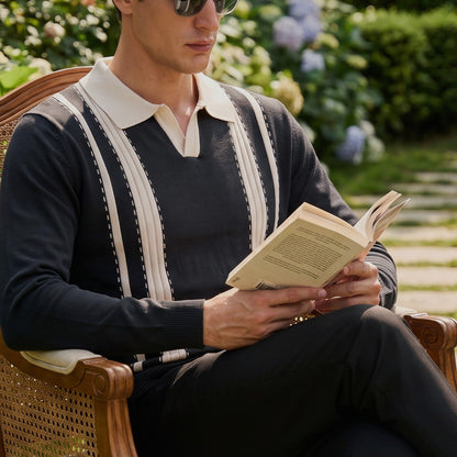 A man wearing aviator sunglasses and a navy knit sweater with cream stripes sits in a wooden garden chair, reading a book.