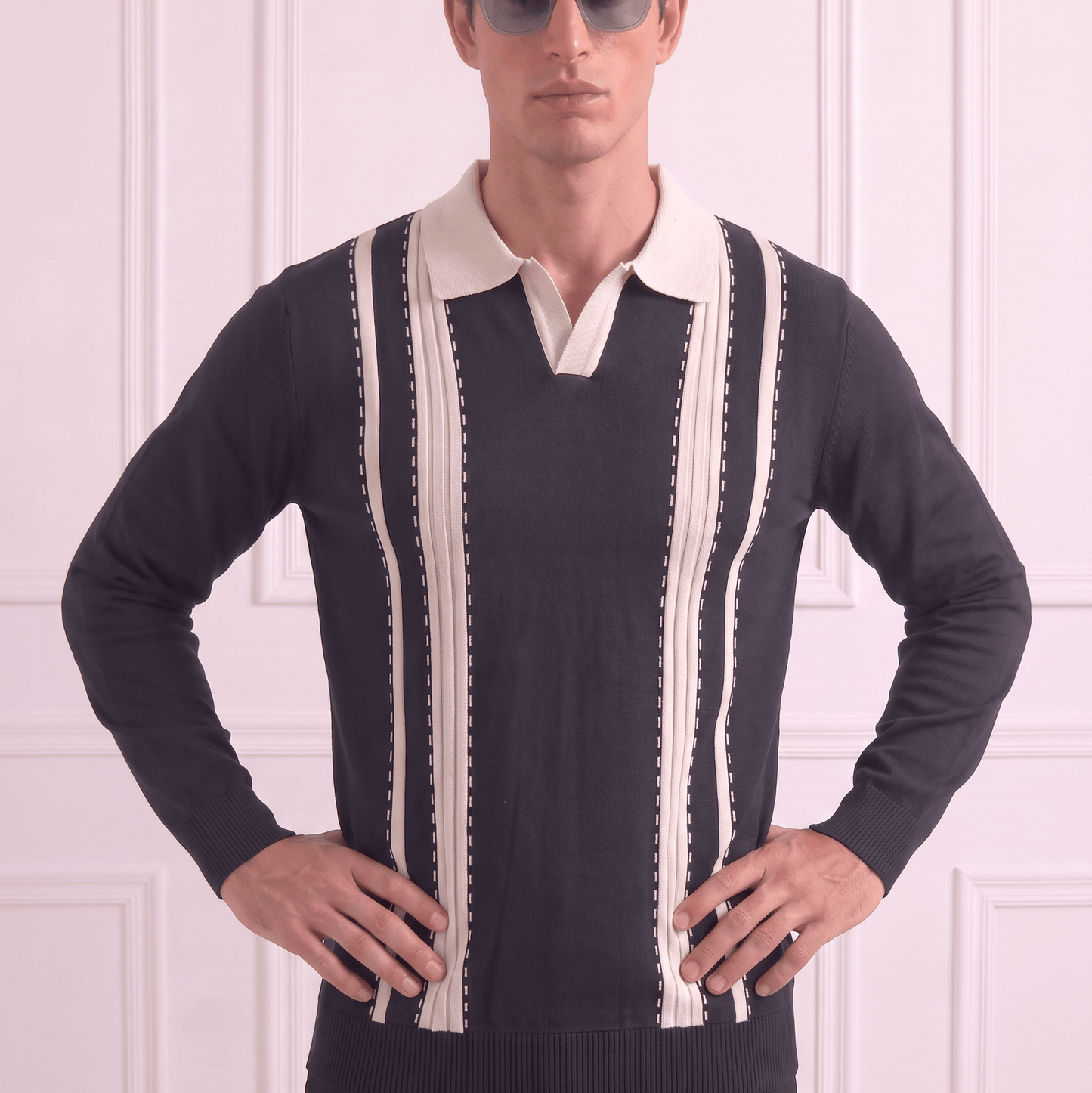 A man wearing a black knit sweater with cream vertical stripes and a cream polo collar, styled with dark sunglasses and black trousers, standing against a white paneled wall.