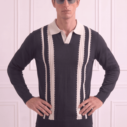 A man wearing a black knit sweater with cream vertical stripes and a cream polo collar, styled with dark sunglasses and black trousers, standing against a white paneled wall.