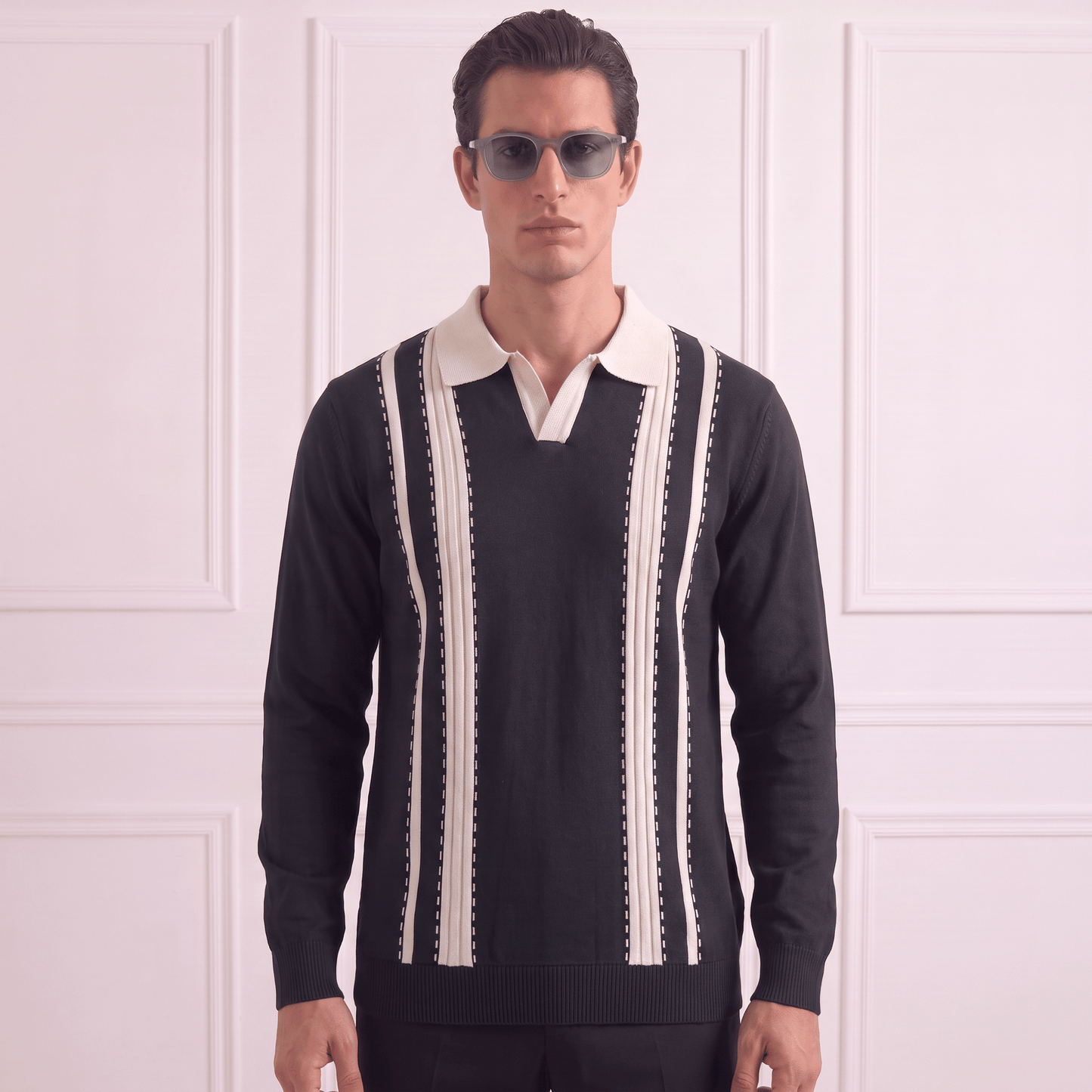 A man wearing a black knit polo sweater with cream vertical stripes and a cream collar, paired with dark sunglasses, standing against a white paneled wall.