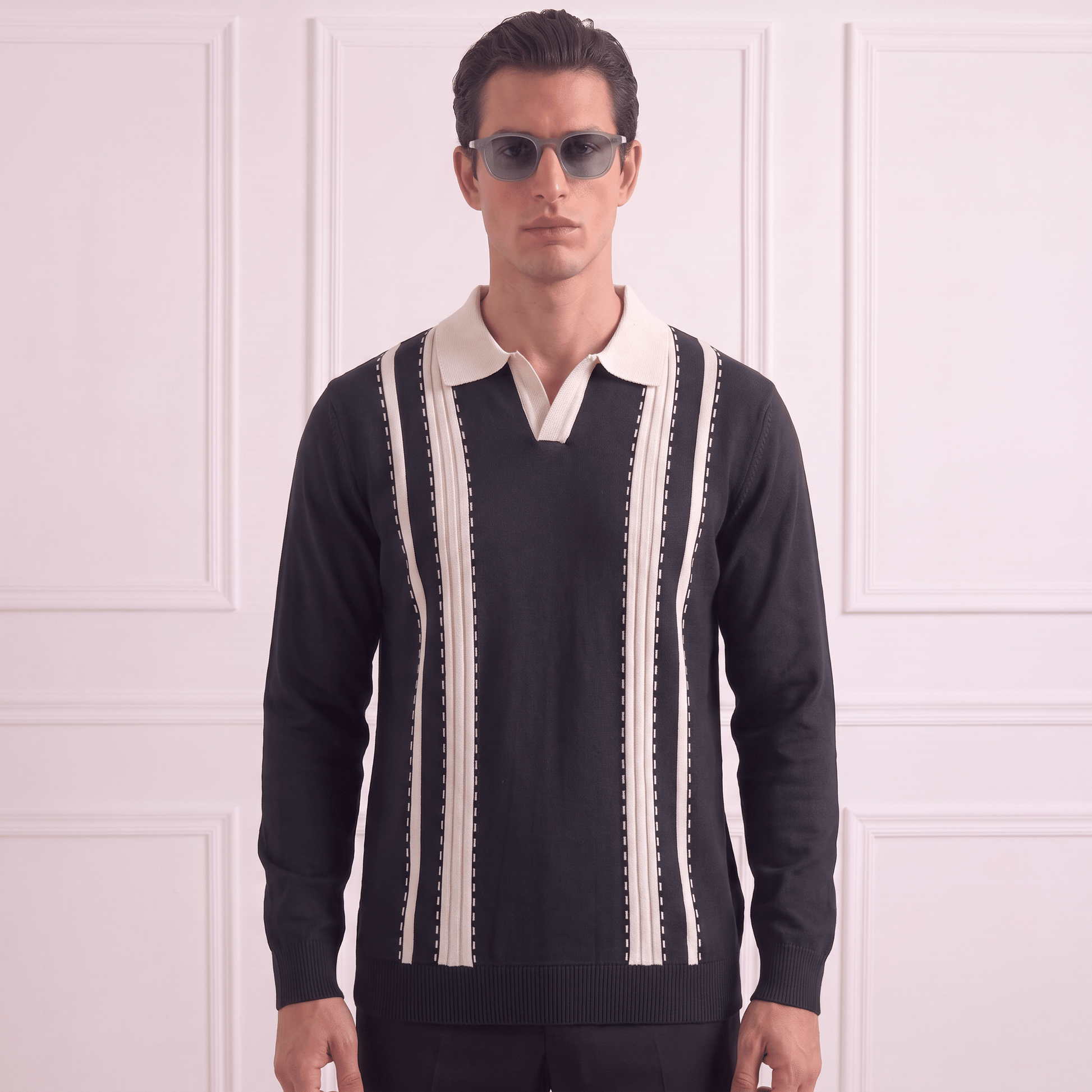 A man wearing a black knit polo sweater with cream vertical stripes and a cream collar, paired with dark sunglasses, standing against a white paneled wall.