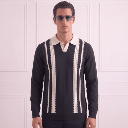 A man wearing a black knit polo sweater with cream vertical stripes and a cream collar, paired with dark sunglasses, standing against a white paneled wall.