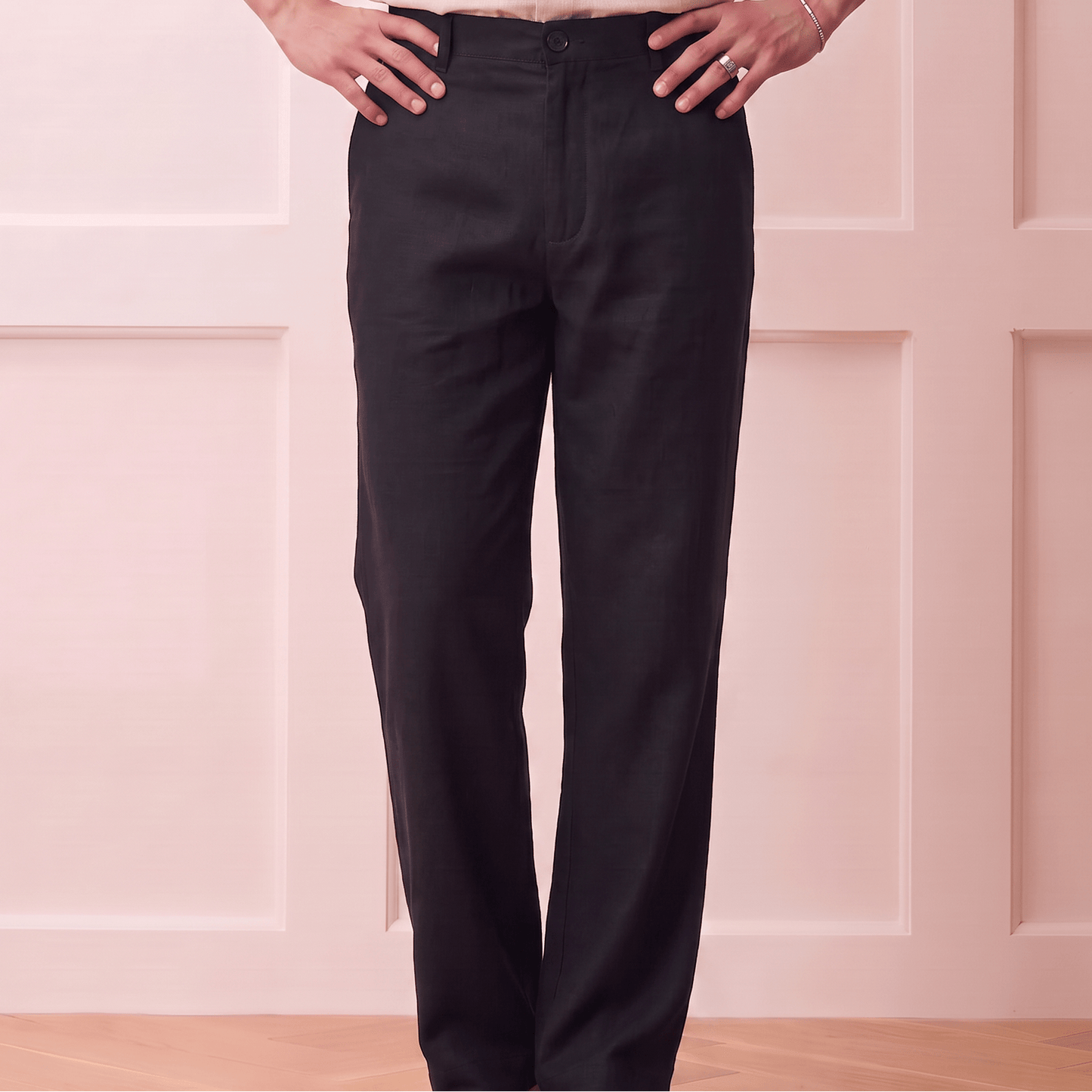 A person wearing black straight-leg trousers and a light beige linen button-up shirt, standing with hands on hips against a pale pink paneled wall.