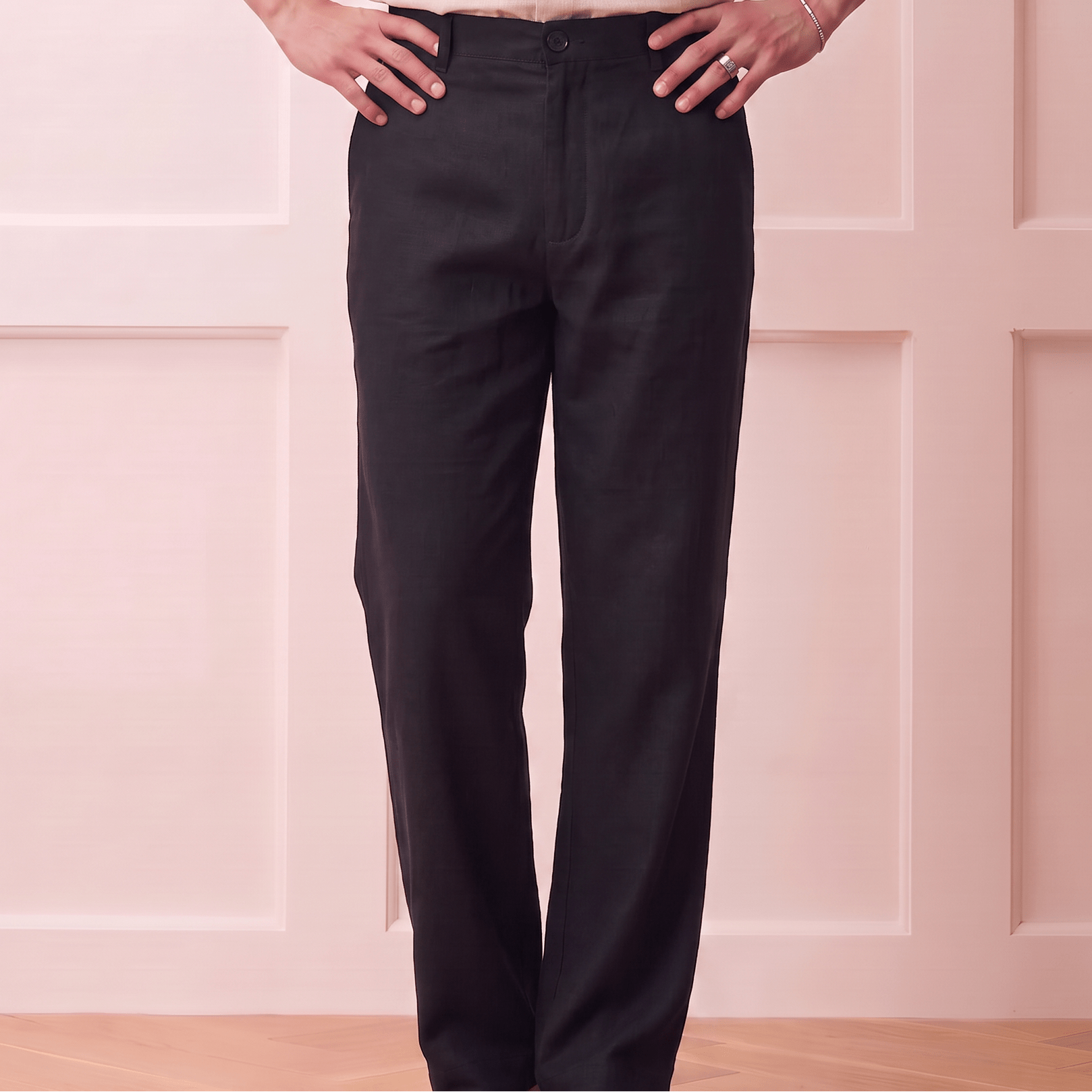 A person wearing black straight-leg trousers and a light beige linen button-up shirt, standing with hands on hips against a pale pink paneled wall.