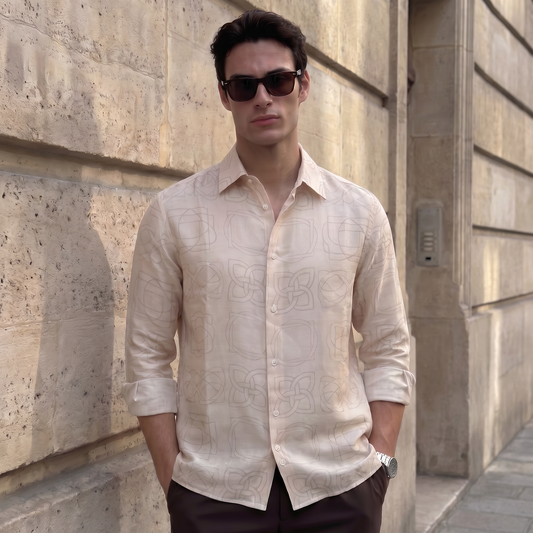 A man wearing a light beige patterned linen shirt and dark sunglasses, standing against a beige stone wall.