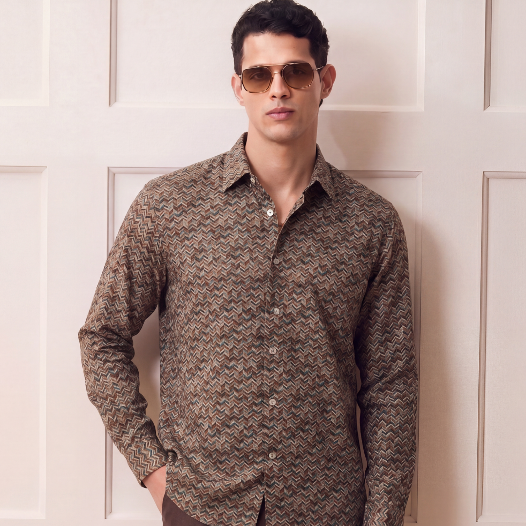A man wearing a brown and beige patterned long-sleeve button-down shirt with dark brown trousers, standing in front of a cream-colored paneled door.