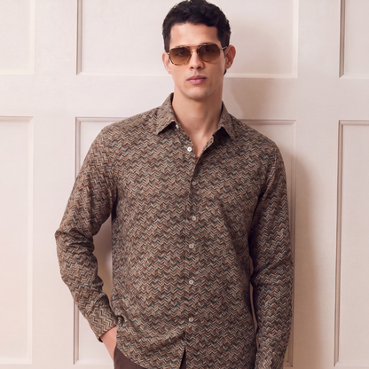 A man wearing a brown and beige patterned long-sleeve button-down shirt with dark brown trousers, standing in front of a cream-colored paneled door.