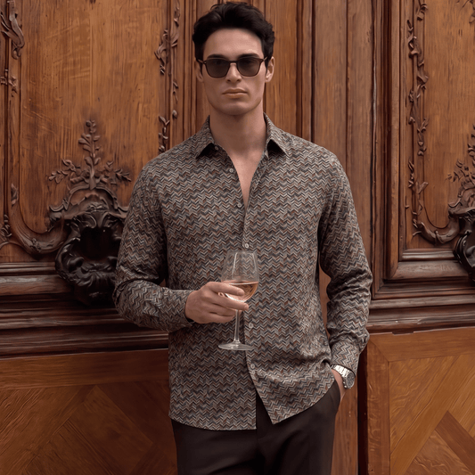A man wearing a brown and beige patterned long-sleeve button-down shirt, dark brown trousers, and sunglasses, holding a glass of white wine in front of a carved wooden door.