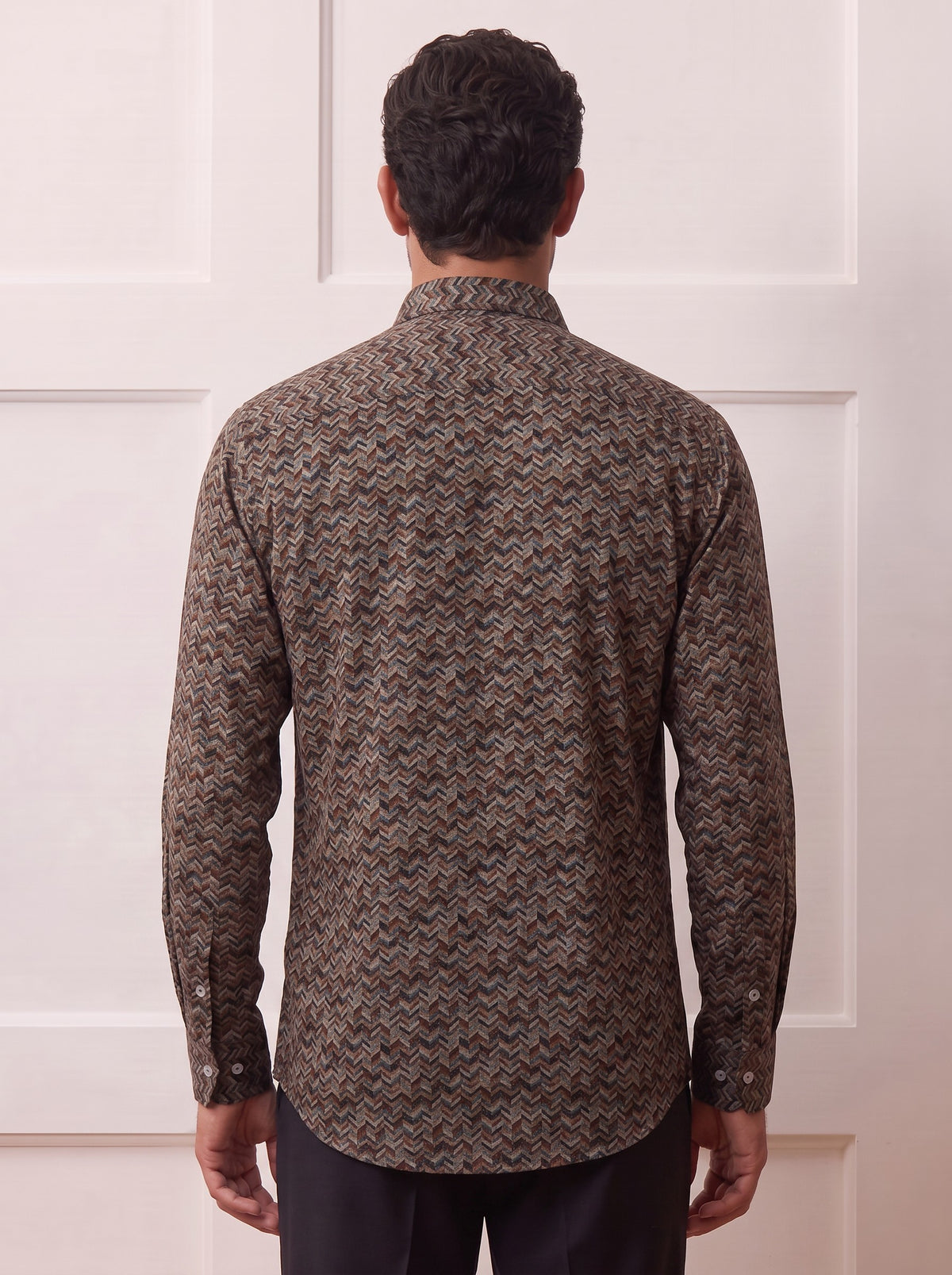 A man viewed from behind wearing a long-sleeve button-up shirt with a brown and beige herringbone pattern, paired with dark trousers, standing against a white paneled wall.