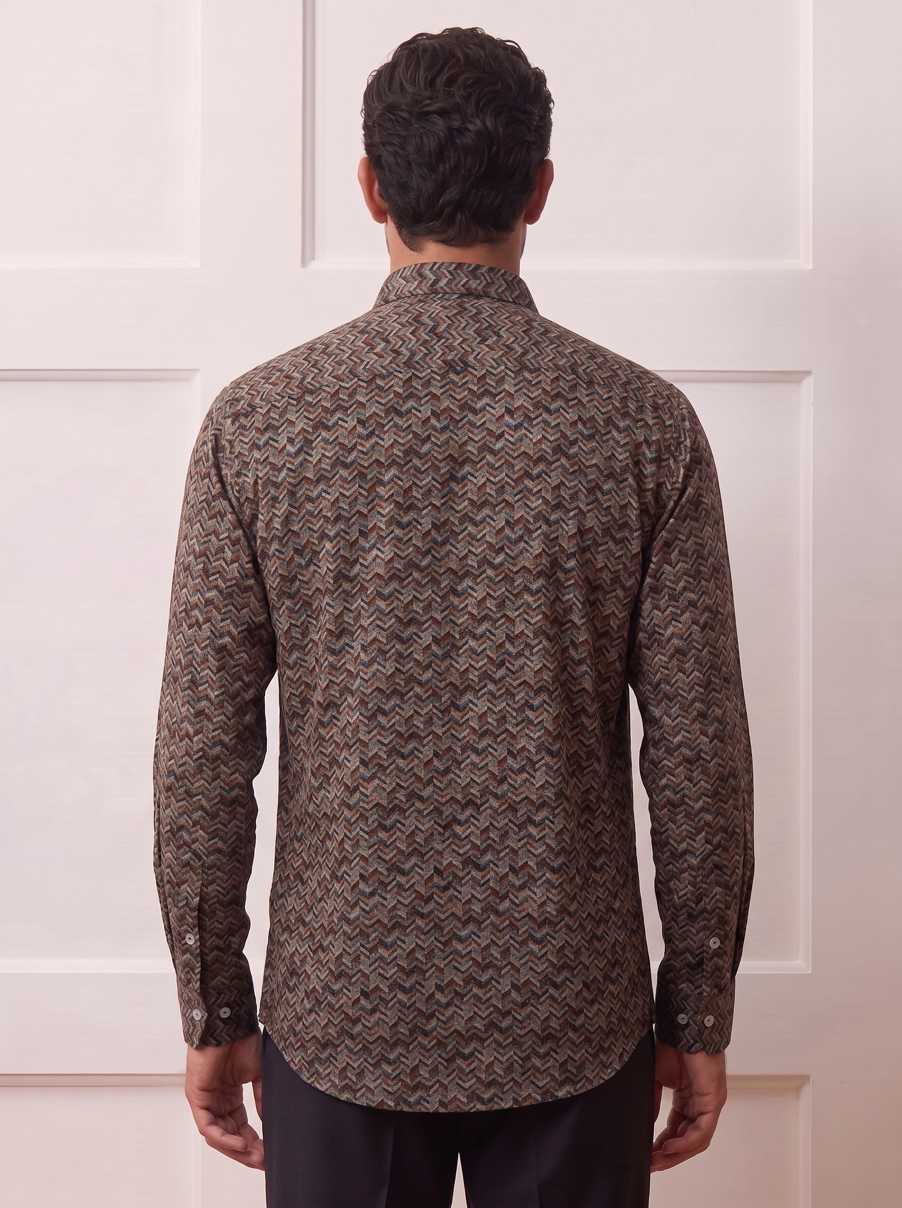 A man viewed from behind wearing a long-sleeve button-up shirt with a brown and beige herringbone pattern, paired with dark trousers, standing against a white paneled wall.