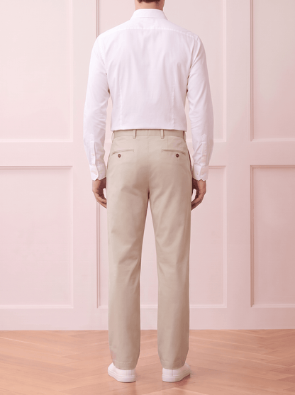 A man viewed from behind wearing a Beige long-sleeve button-up shirt and light beige trousers, standing against a white textured wall.