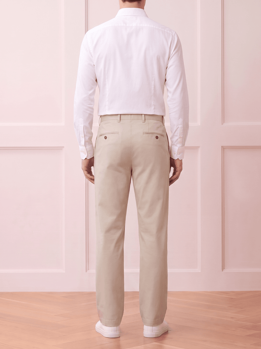 A man viewed from behind wearing a Beige long-sleeve button-up shirt and light beige trousers, standing against a white textured wall.