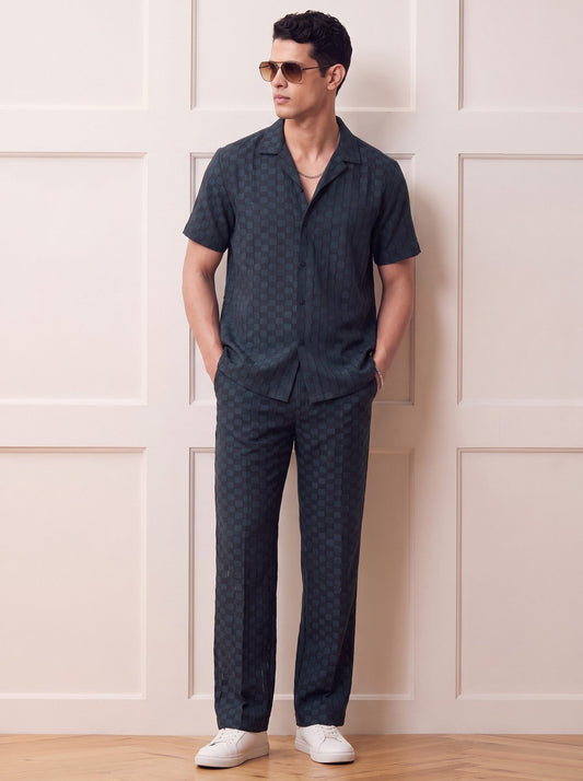 A man wearing a matching dark patterned short-sleeve button-up shirt and trousers set, styled with white sneakers and sunglasses, standing against a cream paneled wall.