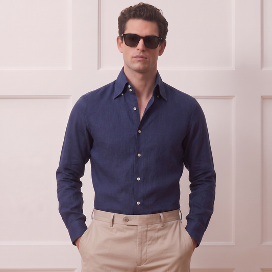 A man wearing a dark navy blue long-sleeve button-down shirt and beige trousers, standing against a light cream paneled wall.