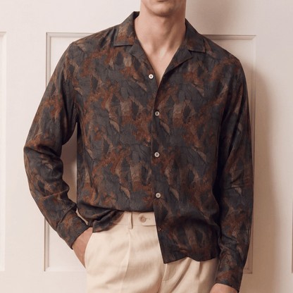 A man wearing a dark brown and black abstract patterned button-down shirt with beige trousers, standing in front of a cream-colored paneled door.