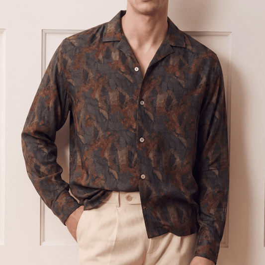 A man wearing a dark brown and black abstract patterned button-down shirt with beige trousers, standing in front of a cream-colored paneled door.