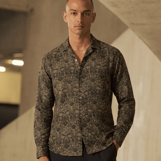 A man wearing a long-sleeve button-up shirt with an intricate dark olive brown and black damask pattern, paired with black trousers, standing in a modern concrete hallway.