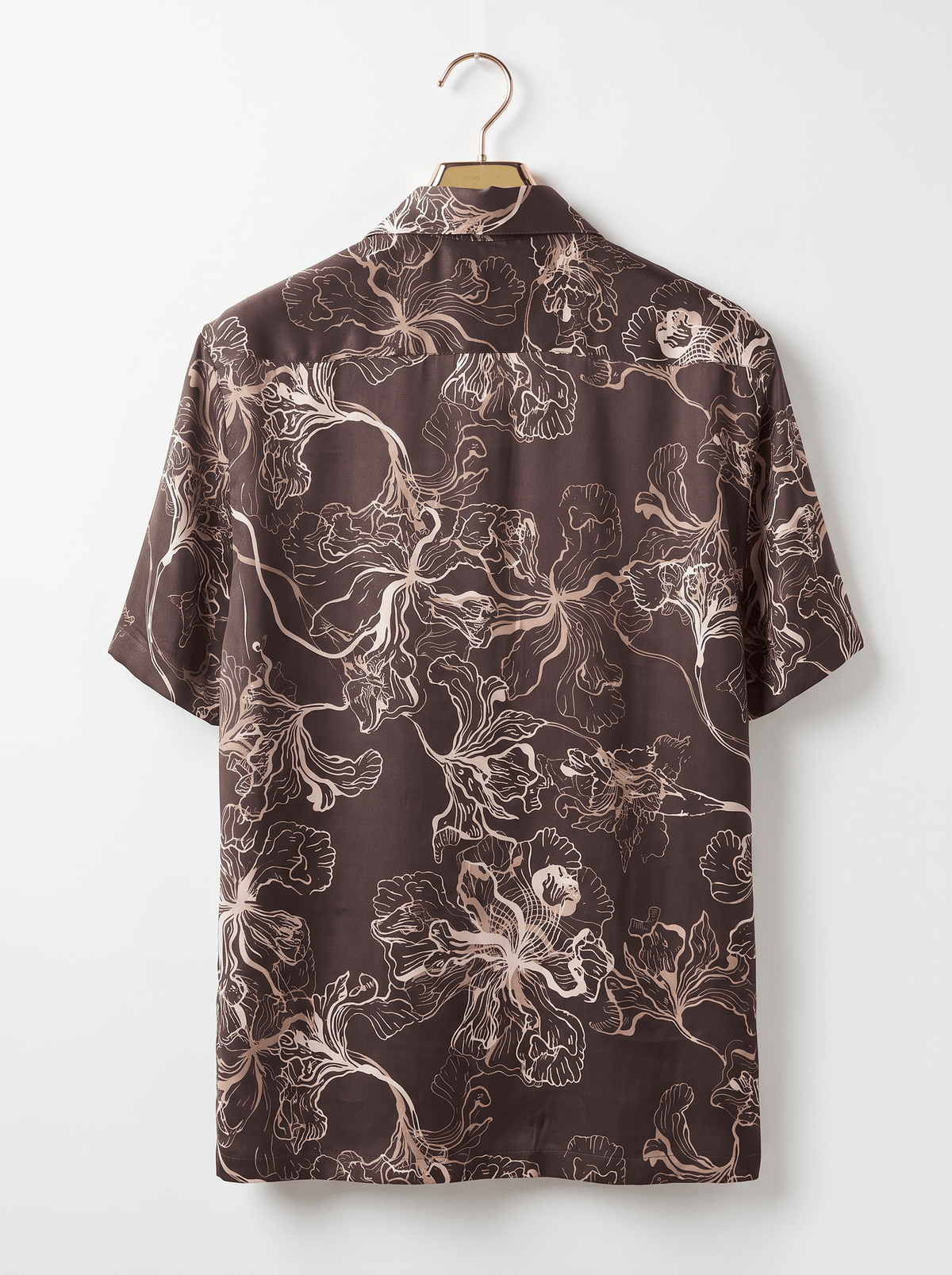 A short-sleeve button-up shirt with an abstract floral line pattern in beige and white on a dark brown background, hanging on a wooden hanger against a plain white wall.