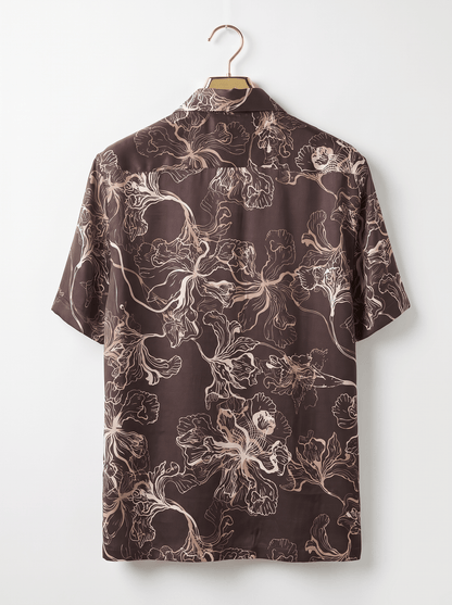 A short-sleeve button-up shirt with an abstract floral line pattern in beige and white on a dark brown background, hanging on a wooden hanger against a plain white wall.