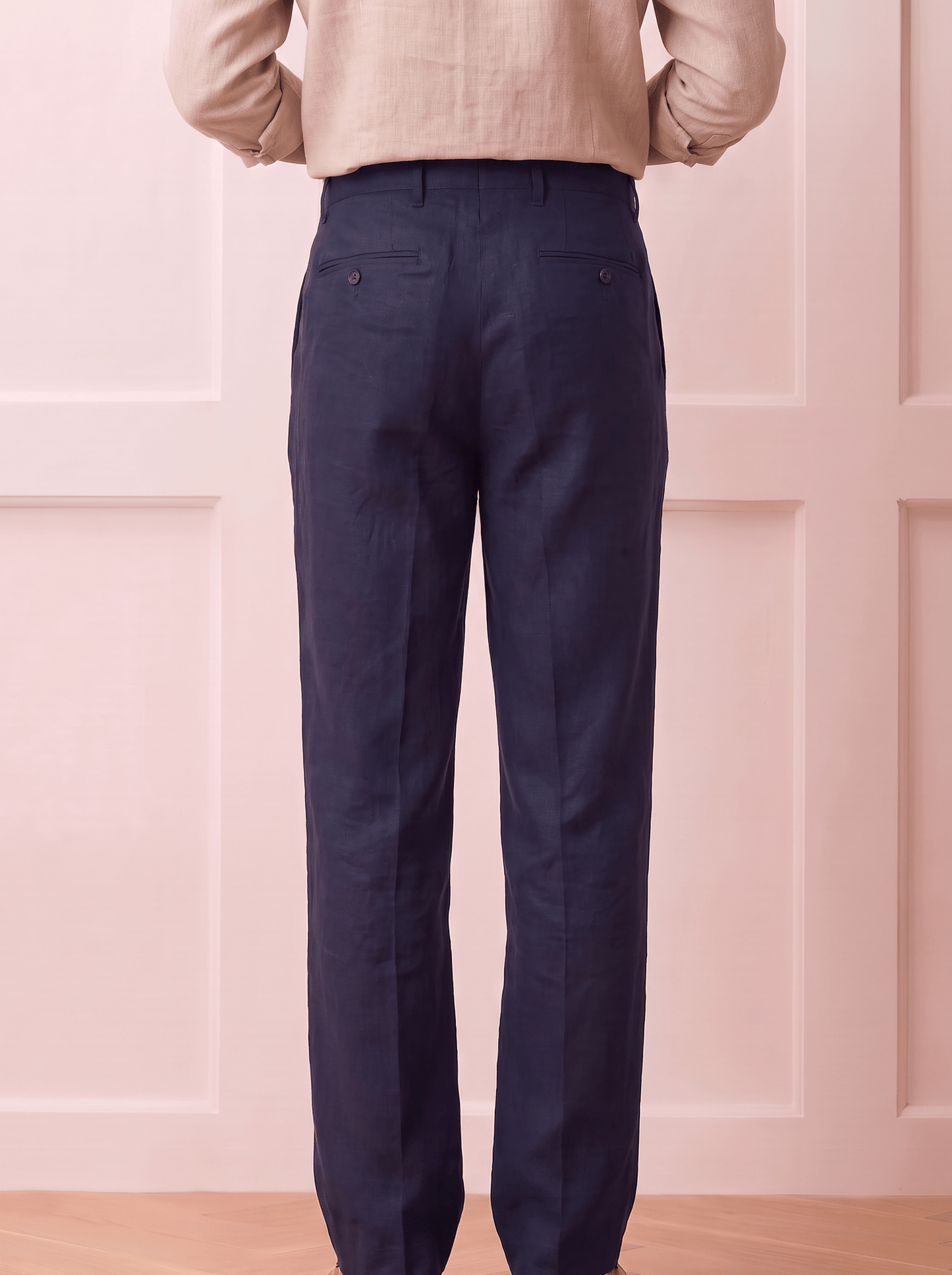 A person shown from the back wearing dark Blue trousers and a light beige top, standing against a pale pink paneled wall.