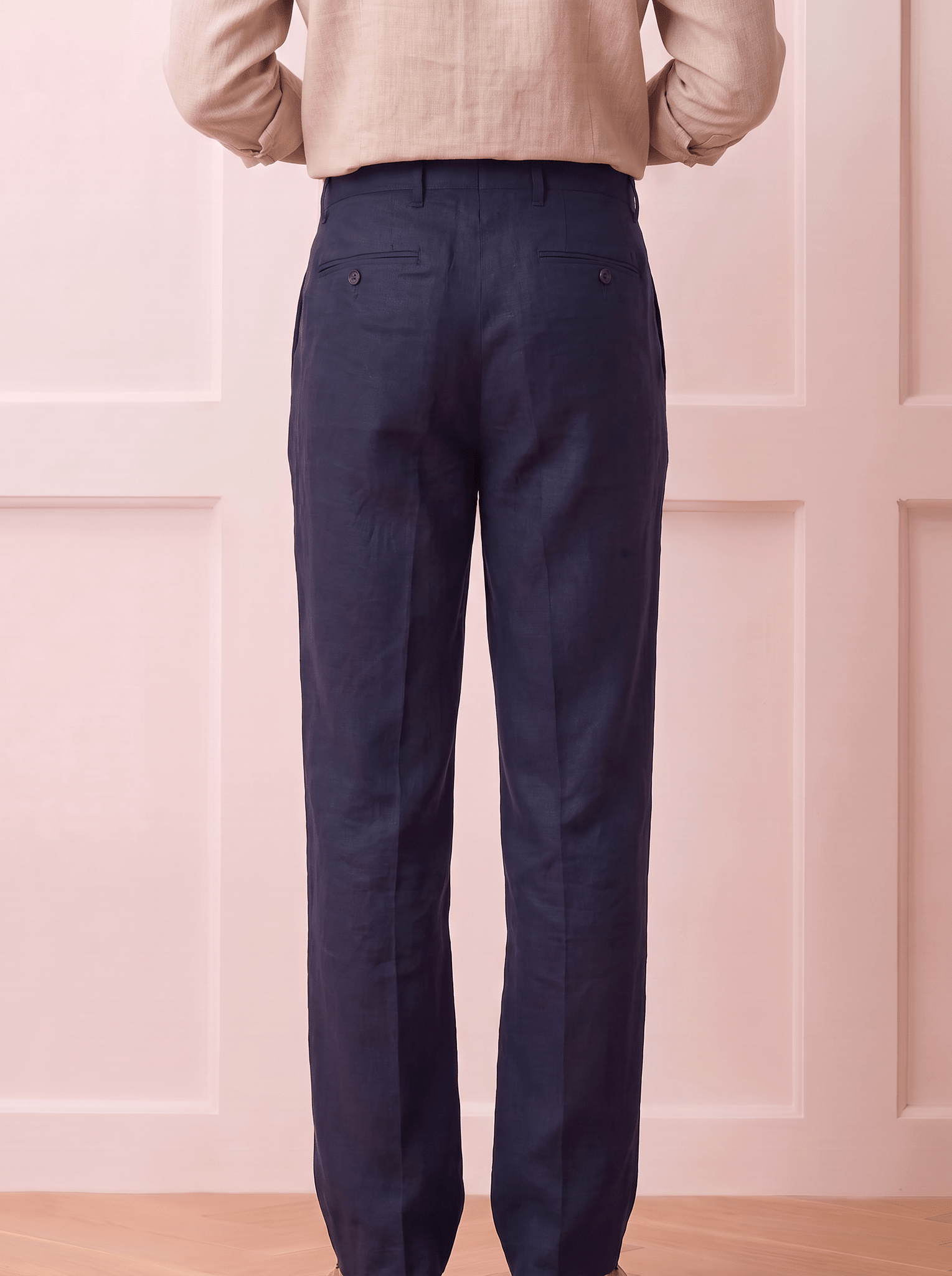 A person shown from the back wearing dark Blue trousers and a light beige top, standing against a pale pink paneled wall.