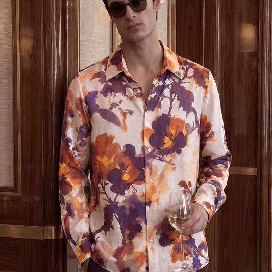 A man wearing a long-sleeve button-up shirt with a vibrant orange, purple, and yellow floral print on a cream background, styled with dark sunglasses and holding a glass of white wine in front of a dark wood-paneled wall.
