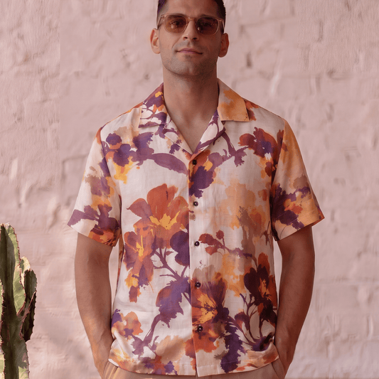 A man wearing a short-sleeve button-up shirt with a vibrant orange, purple, and yellow floral print, paired with beige trousers, standing against a textured light pink wall.