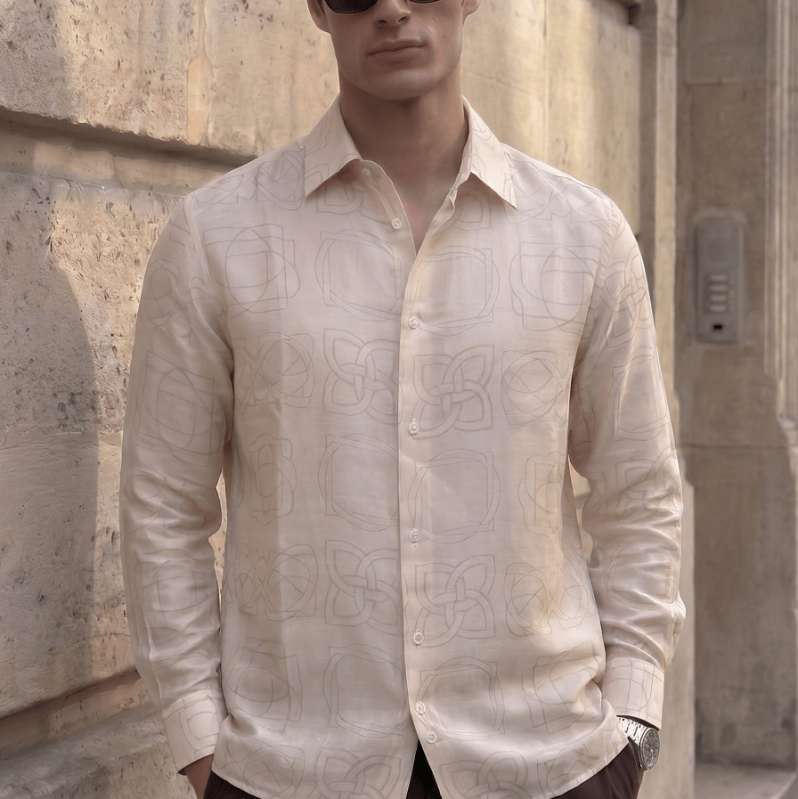 A man wearing a light beige long-sleeve button-up shirt with a subtle tonal geometric pattern, dark sunglasses, and dark trousers, standing against a textured stone wall.