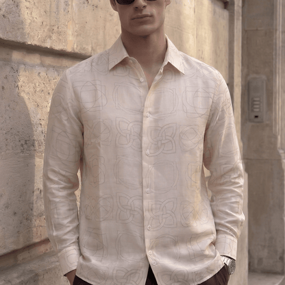A man wearing a light beige long-sleeve button-up shirt with a subtle tonal geometric pattern, dark sunglasses, and dark trousers, standing against a textured stone wall.